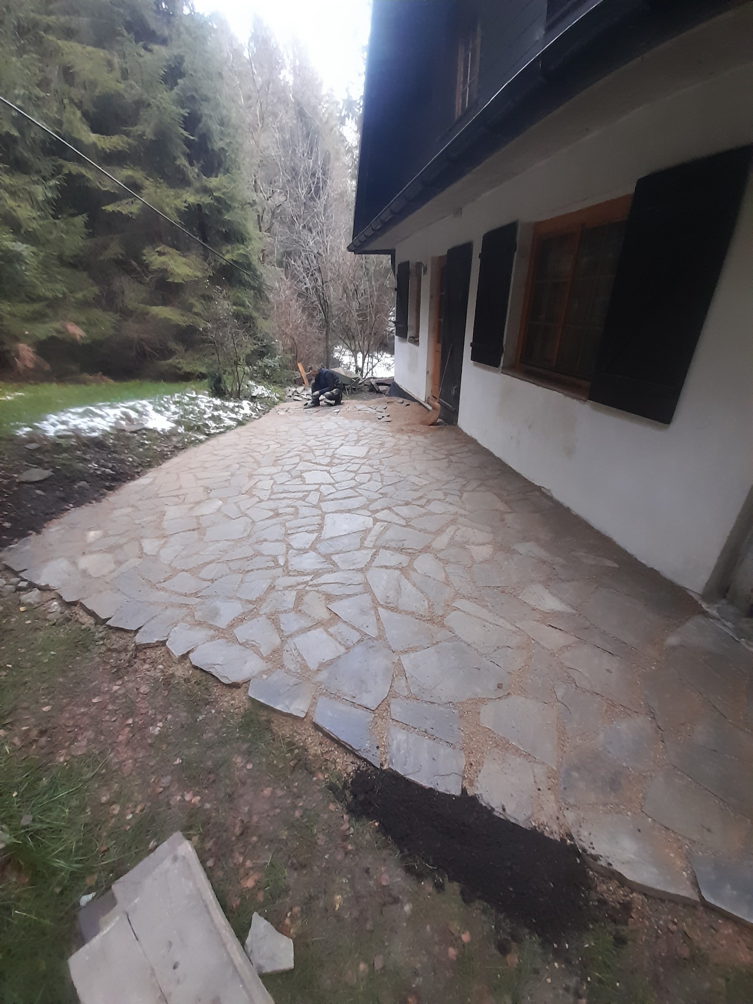 Układanie nieregularnych kamiennych płyt chodnikowych na zewnątrz budynku, widoczny pracownik w oddali, teren częściowo pokryty śniegiem.