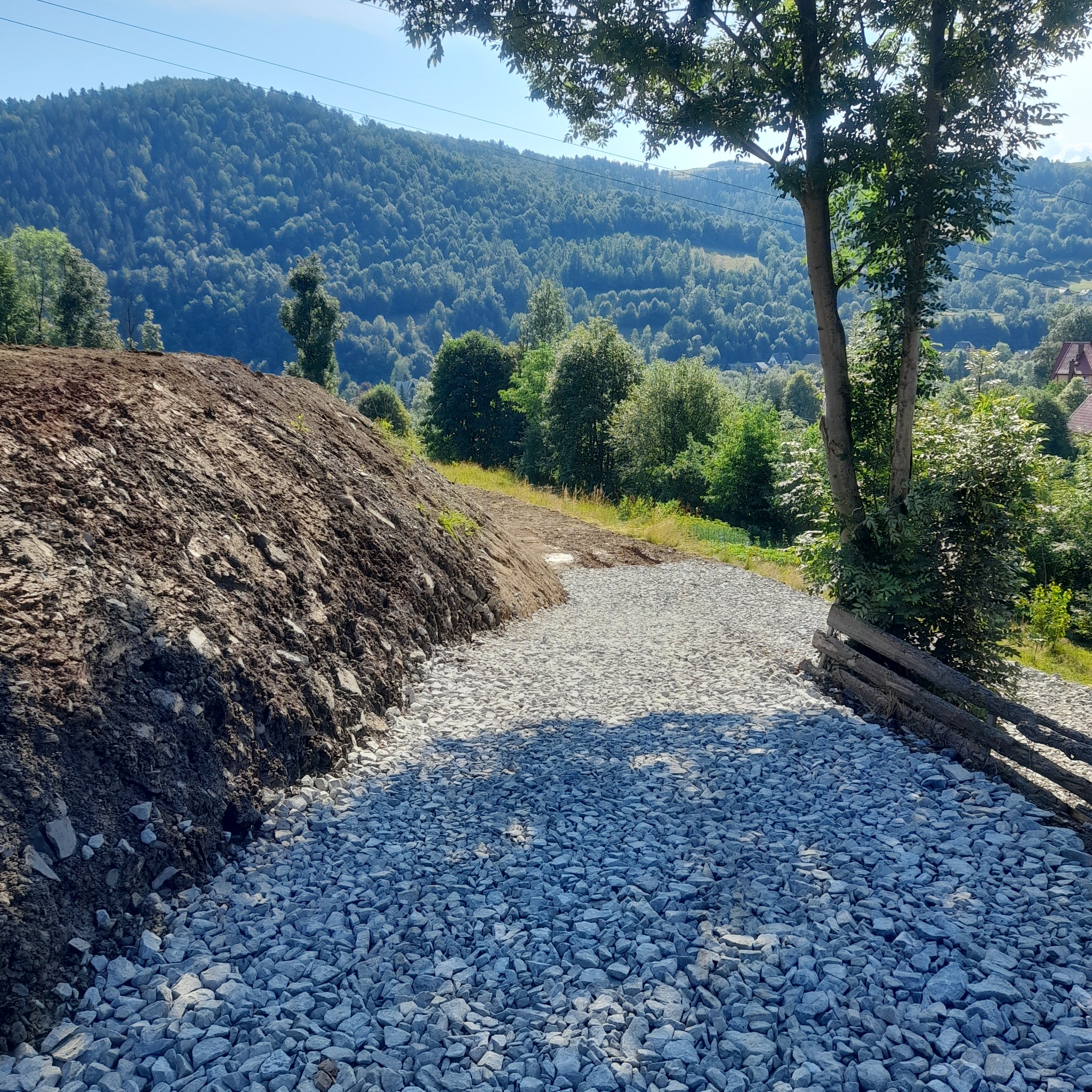 Świeżo utwardzona droga z szarego kruszywa, obok nasyp ziemny z widokiem na zalesione wzgórza w tle, obrzeże z drewnianych bali przy drzewie.