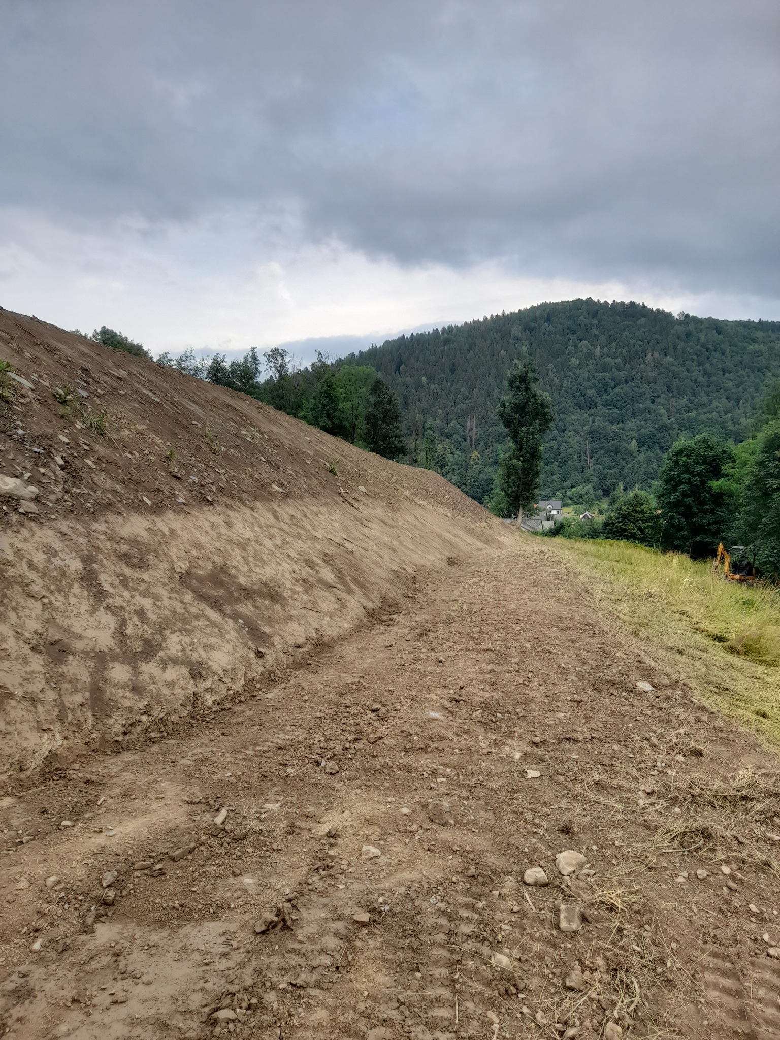 Ukształtowany, piaszczysty teren na zboczu wzgórza z widocznym w oddali lasem i pochmurnym niebem, mała koparka po prawej stronie.