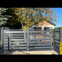 Nowoczesna brama wjazdowa z poziomych profili w kolorze grafitowym, częściowo otwarta, z widocznym logo producenta, na tle domu z ogrodem w jesiennej scenerii.