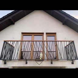 Ślusarstwo Nosidlak Damian - Balkon z czarną, geometryczną balustradą z ukośnych prętów i drewnianym poręczą, widok z dołu na tle jasnej elewacji domu z dwoma oknami i logiem firmy na balkonie.