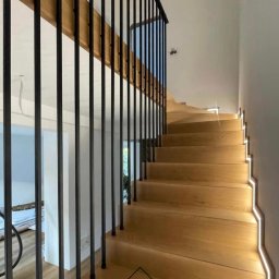 Ślusarstwo Nosidlak Damian - Drewniane schody z czarną, metalową balustradą i subtelnym oświetleniem LED wzdłuż stopni.