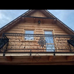 Ślusarstwo Nosidlak Damian - Kuta balustrada balkonowa z motywem roślinnym i ozdobnym medalionem na drewnianym domu z dwoma oknami, widok z dołu, logo firmy na balustradzie.