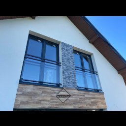 Ślusarstwo Nosidlak Damian - Elewacja domu z dwoma oknami balkonowymi zabezpieczonymi nowoczesnymi balustradami z czarnymi ramami i szklanymi panelami, z widocznym logo firmy na elewacji.