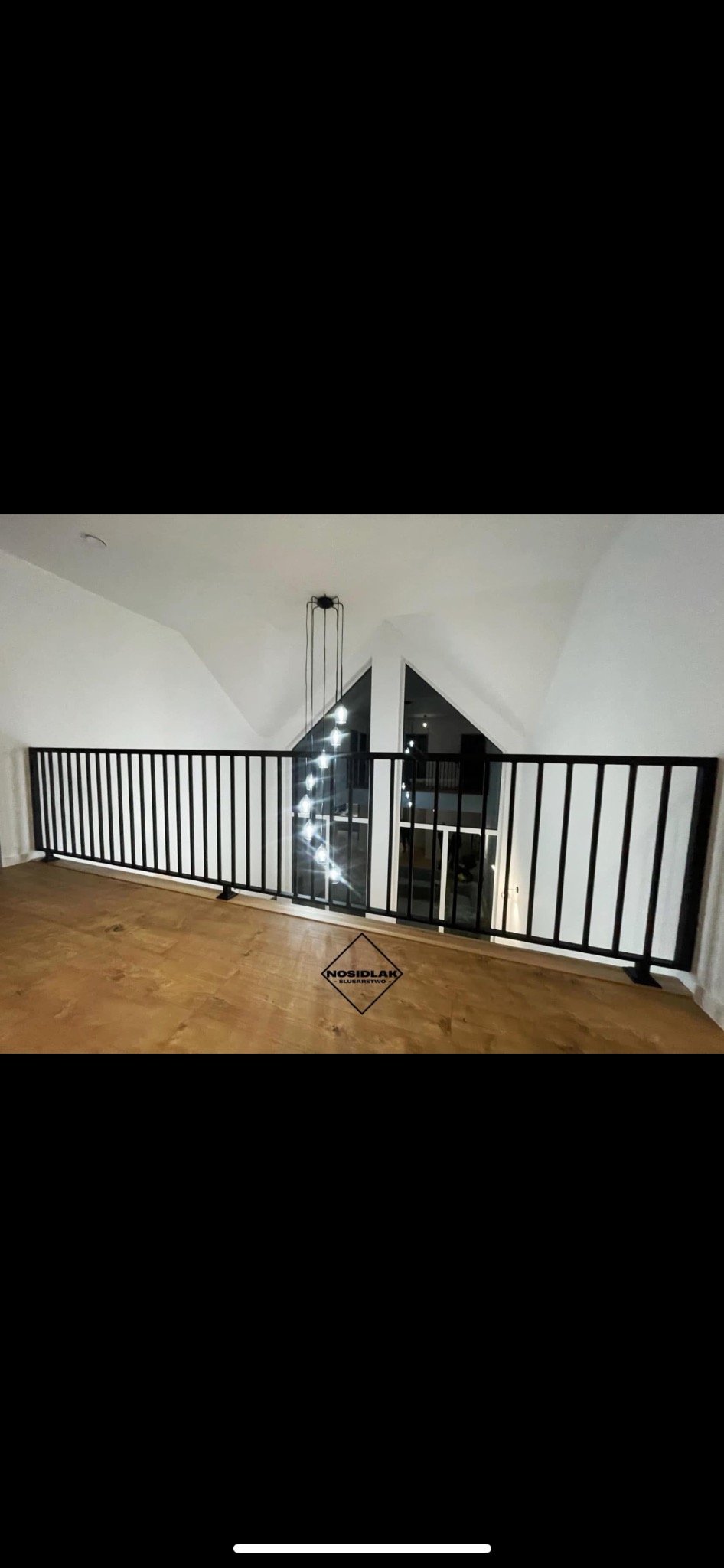 Wewnętrzna balustrada z czarnych, pionowych prętów na drewnianej podłodze, widok z góry, w tle okno i wisząca lampa, logo firmy na pierwszym planie.