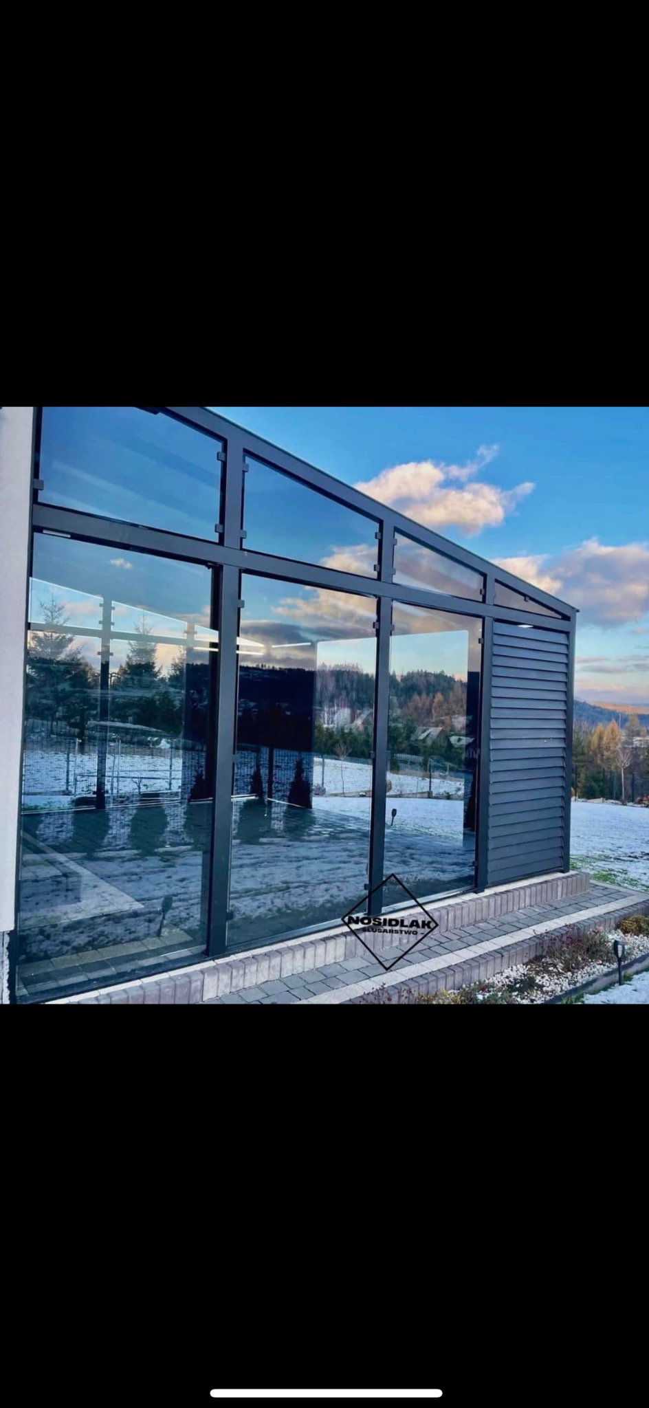 Nowoczesna pergola z metalową konstrukcją i szklanymi ścianami, widok z zewnątrz na tle zimowego krajobrazu z resztkami śniegu i błękitnym niebem.