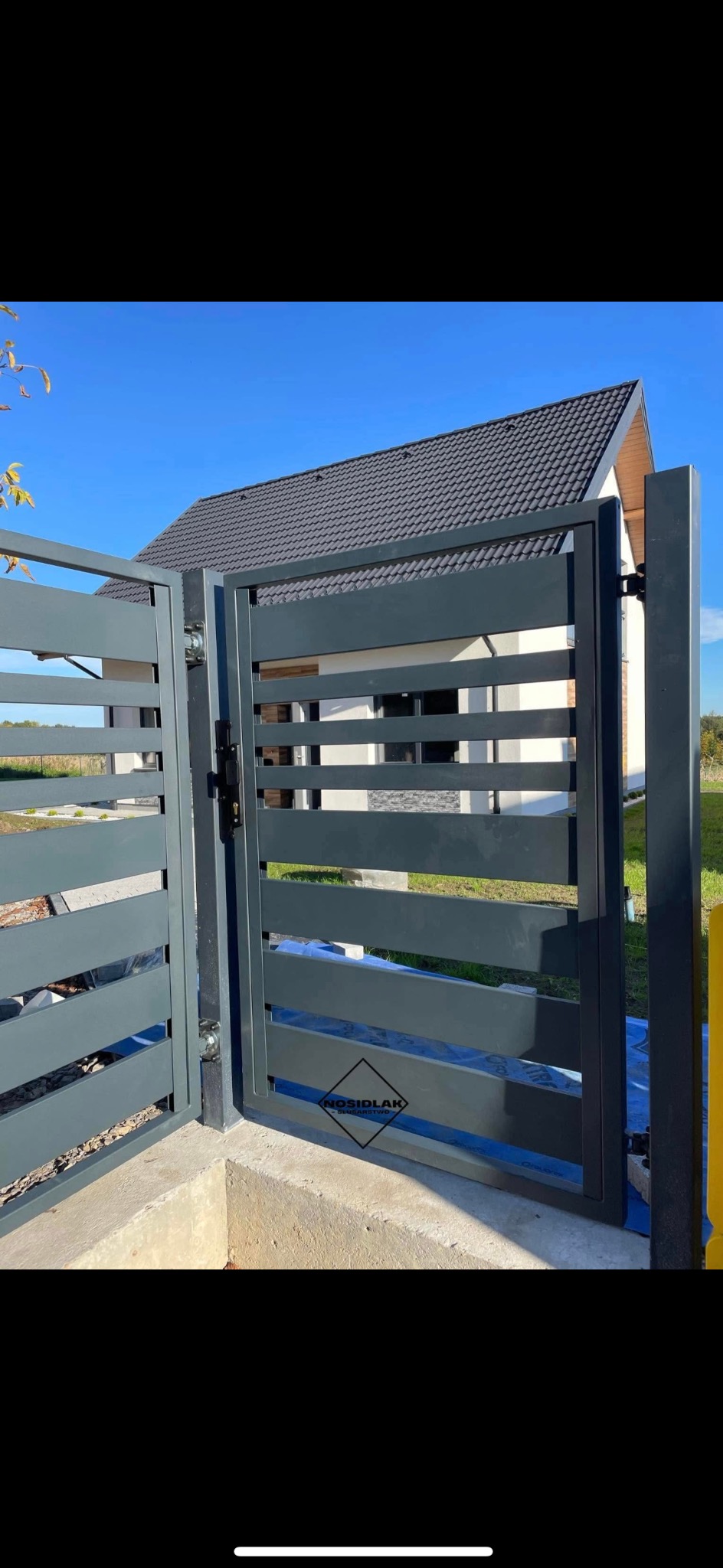 Nowoczesna, grafitowa brama wjazdowa z poziomymi panelami, częściowo otwarta, na tle domu z ciemnym dachem i błękitnego nieba.