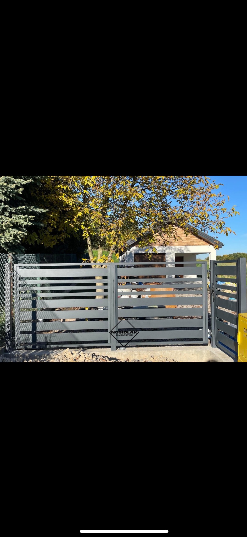 Nowoczesna brama wjazdowa z poziomych profili w kolorze grafitowym, częściowo otwarta, z widocznym logo producenta, na tle domu z ogrodem w jesiennej scenerii.
