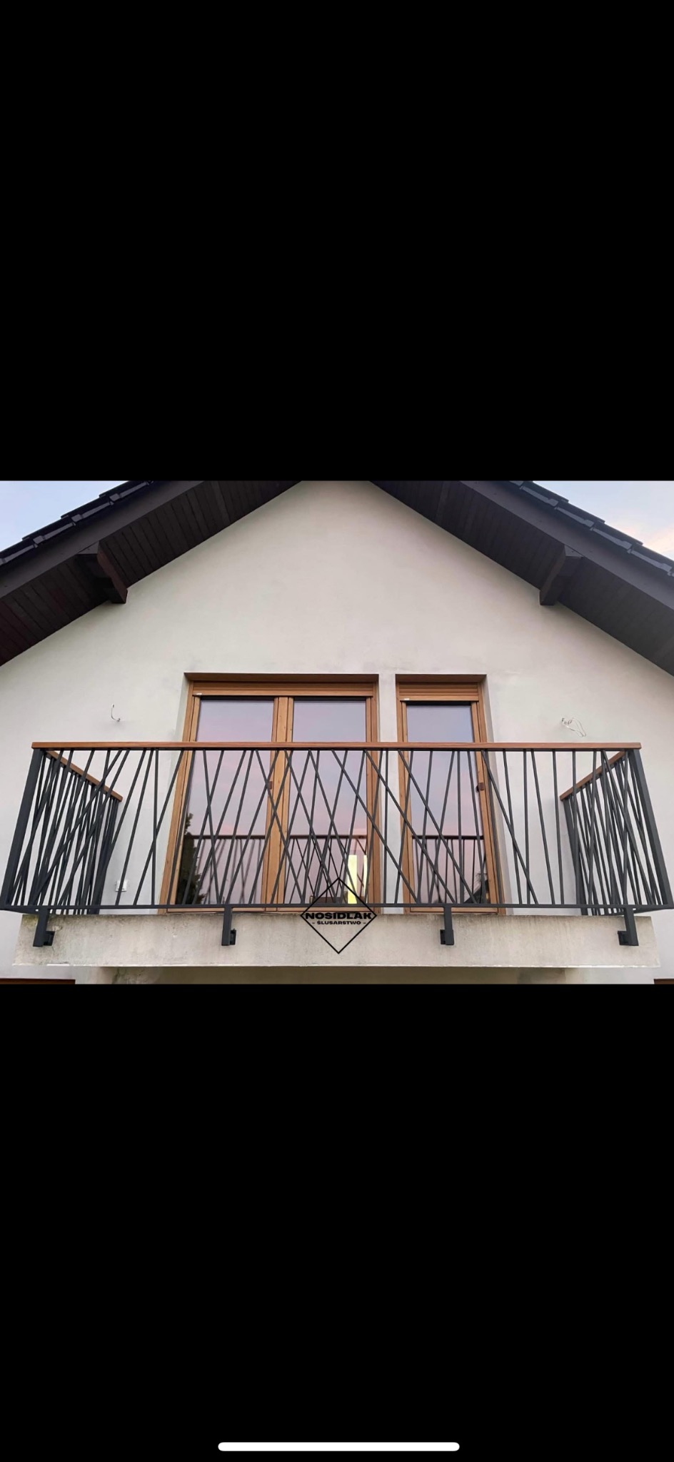 Balkon z czarną, geometryczną balustradą z ukośnych prętów i drewnianym poręczą, widok z dołu na tle jasnej elewacji domu z dwoma oknami i logiem firmy na balkonie.