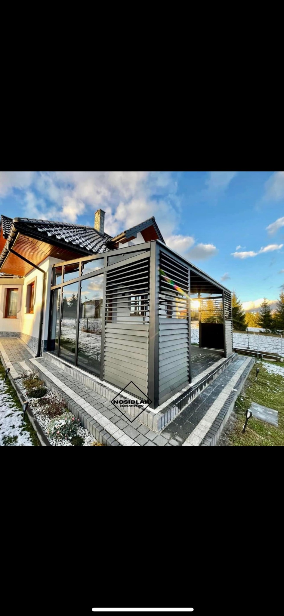 Nowoczesna pergola aluminiowa z regulowanymi lamelami, przylegająca do budynku z dużymi oknami, widok z zewnątrz na tle zimowego krajobrazu i błękitnego nieba.