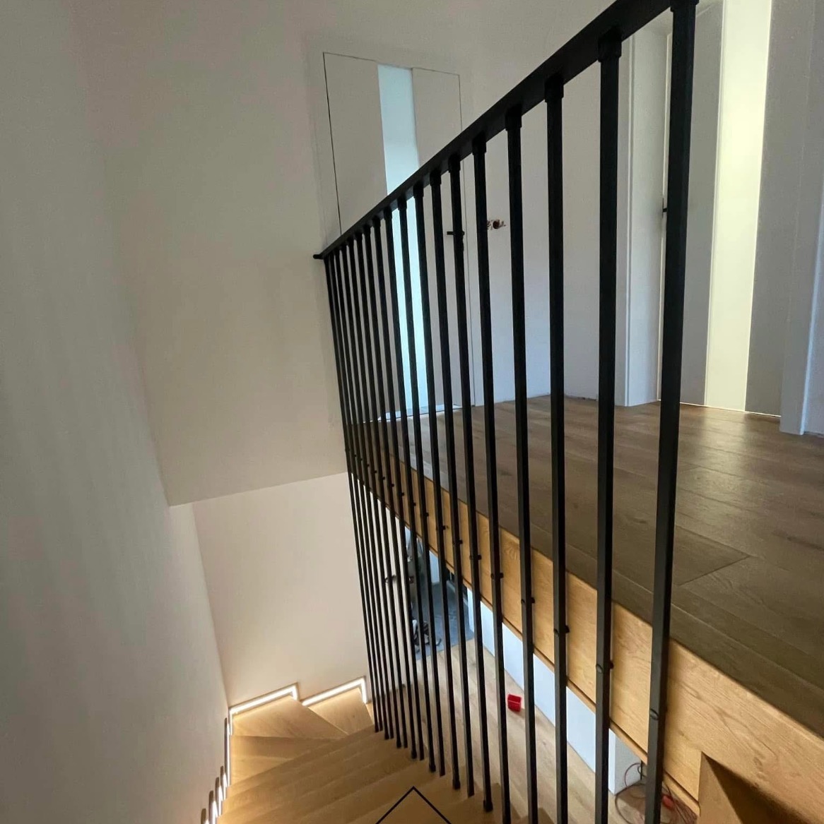 Nowoczesne schody drewniane z czarną, minimalistyczną balustradą z pionowych prętów, oświetlenie LED pod stopniami, widok z góry.