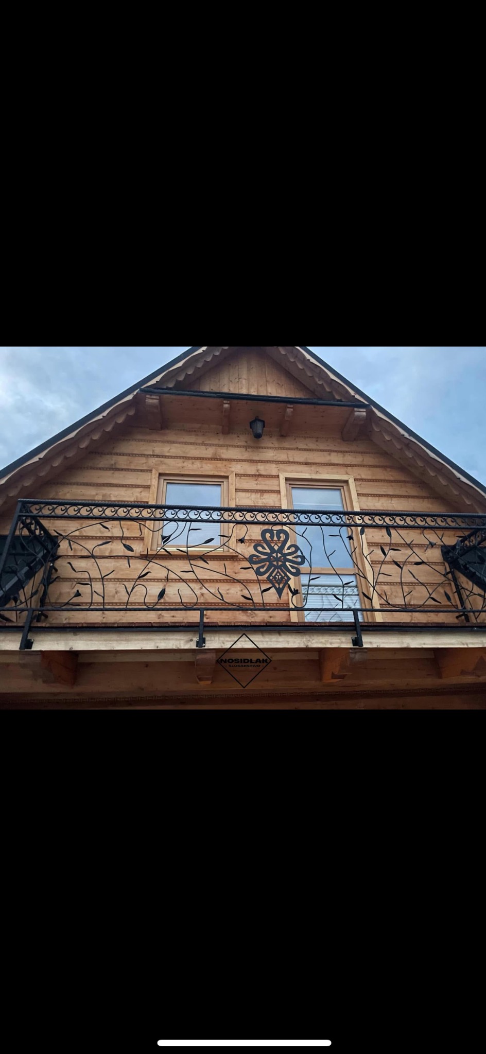 Kuta balustrada balkonowa z motywem roślinnym i ozdobnym medalionem na drewnianym domu z dwoma oknami, widok z dołu, logo firmy na balustradzie.