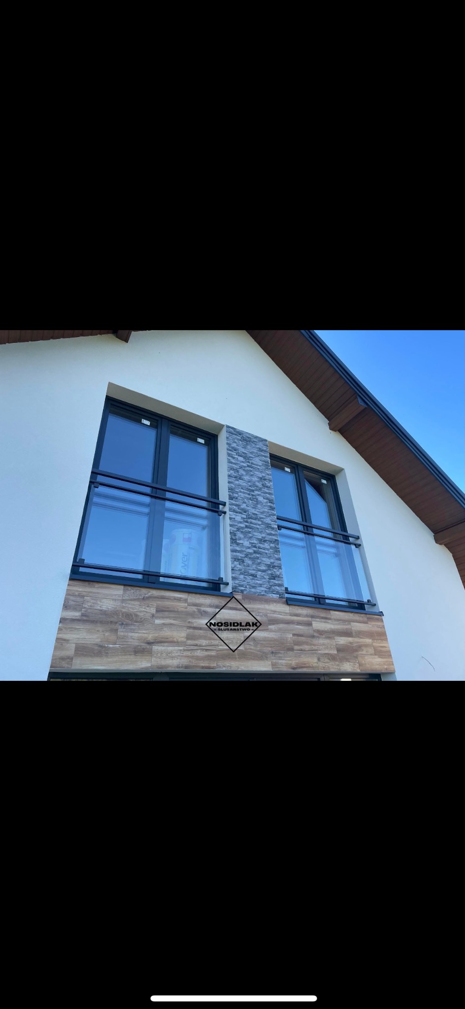 Elewacja domu z dwoma oknami balkonowymi zabezpieczonymi nowoczesnymi balustradami z czarnymi ramami i szklanymi panelami, z widocznym logo firmy na elewacji.