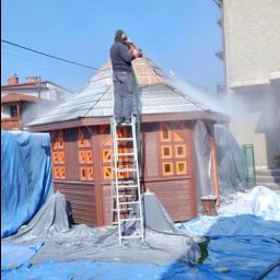 Eco Blast System Walczak Spółka Jawna - Pracownik w kombinezonie ochronnym i słuchawkach, stojący na drabinie, czyści strumieniem wody z węża dach altany, zabezpieczonej folią ochronną, na tle błękitnego nieba i budynków.