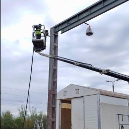 Eco Blast System Walczak Spółka Jawna - Pracownik w podnośniku koszowym czyści metalową konstrukcję przypominającą suwnicę, widoczne koła od wagonów kolejowych, drabina i biały namiot magazynowy w tle.