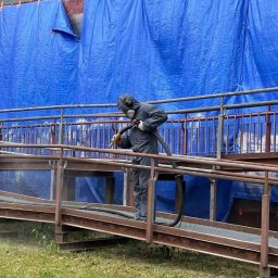 Eco Blast System Walczak Spółka Jawna - Pracownik w kombinezonie ochronnym piaskuje metalową konstrukcję na zewnątrz budynku, zabezpieczonego niebieską plandeką. Widoczny pył i trawa.