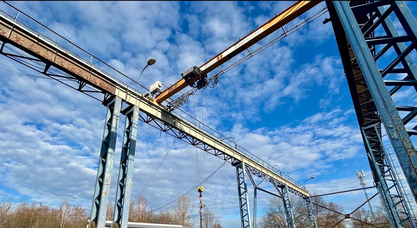 Suwnica bramowa z żółtym wózkiem i napisem 5T, widoczna z dołu na tle błękitnego nieba z chmurami, elementy konstrukcyjne w kolorach niebieskim i pomarańczowym.