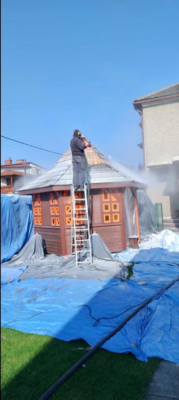 Pracownik w kombinezonie ochronnym i słuchawkach, stojący na drabinie, czyści strumieniem wody z węża dach altany, zabezpieczonej folią ochronną, na tle błękitnego nieba i budynków.
