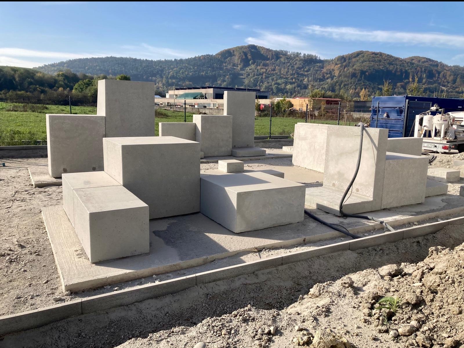 Betonowe bloki ułożone w formie rzeźby plenerowej na placu budowy, z widocznymi przewodami i sprzętem do obróbki powierzchni w tle, na tle zielonych wzgórz.