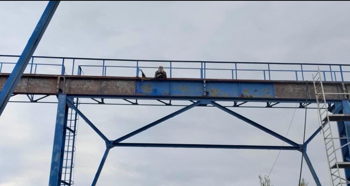 Pracownik na konstrukcji stalowej mostu dźwigowego przygotowuje powierzchnię do malowania, widoczne ślady korozji i częściowo oczyszczona powierzchnia.