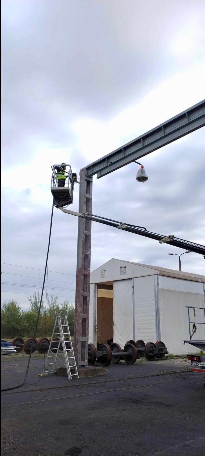 Pracownik w podnośniku koszowym czyści metalową konstrukcję przypominającą suwnicę, widoczne koła od wagonów kolejowych, drabina i biały namiot magazynowy w tle.