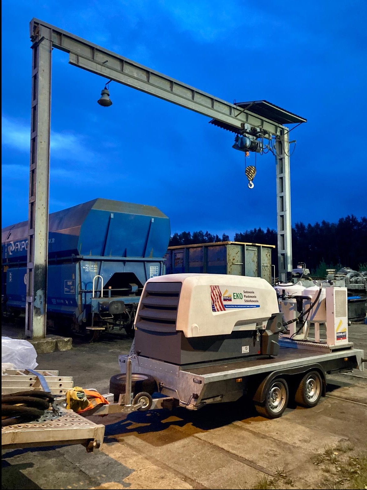 Mobilny zestaw do obróbki strumieniowo-ściernej na przyczepie, z widocznym kompresorem i zbiornikiem, na tle wagonów kolejowych i suwnicy bramowej.