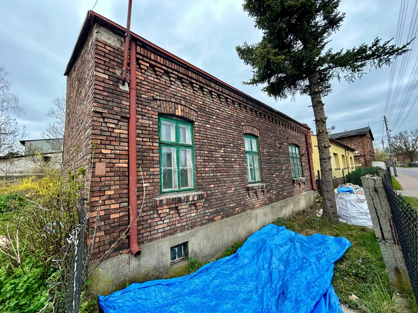 Ceglany budynek z zielonymi oknami, zabezpieczony niebieską plandeką. Widoczny fragment drzewa i sąsiednie budynki. Ujęcie z dołu, podkreślające bryłę budynku.
