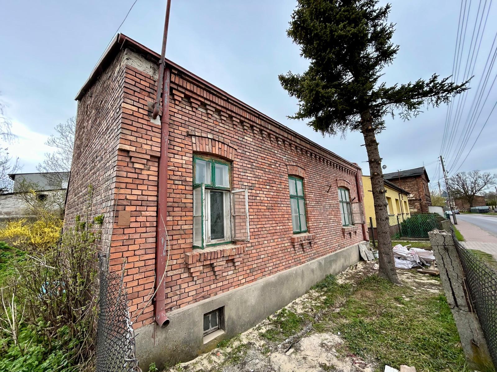 Ceglany budynek z zielonymi ramami okiennymi, widoczny z niskiej perspektywy, z fragmentem drzewa po prawej stronie. Część okna jest otwarta.