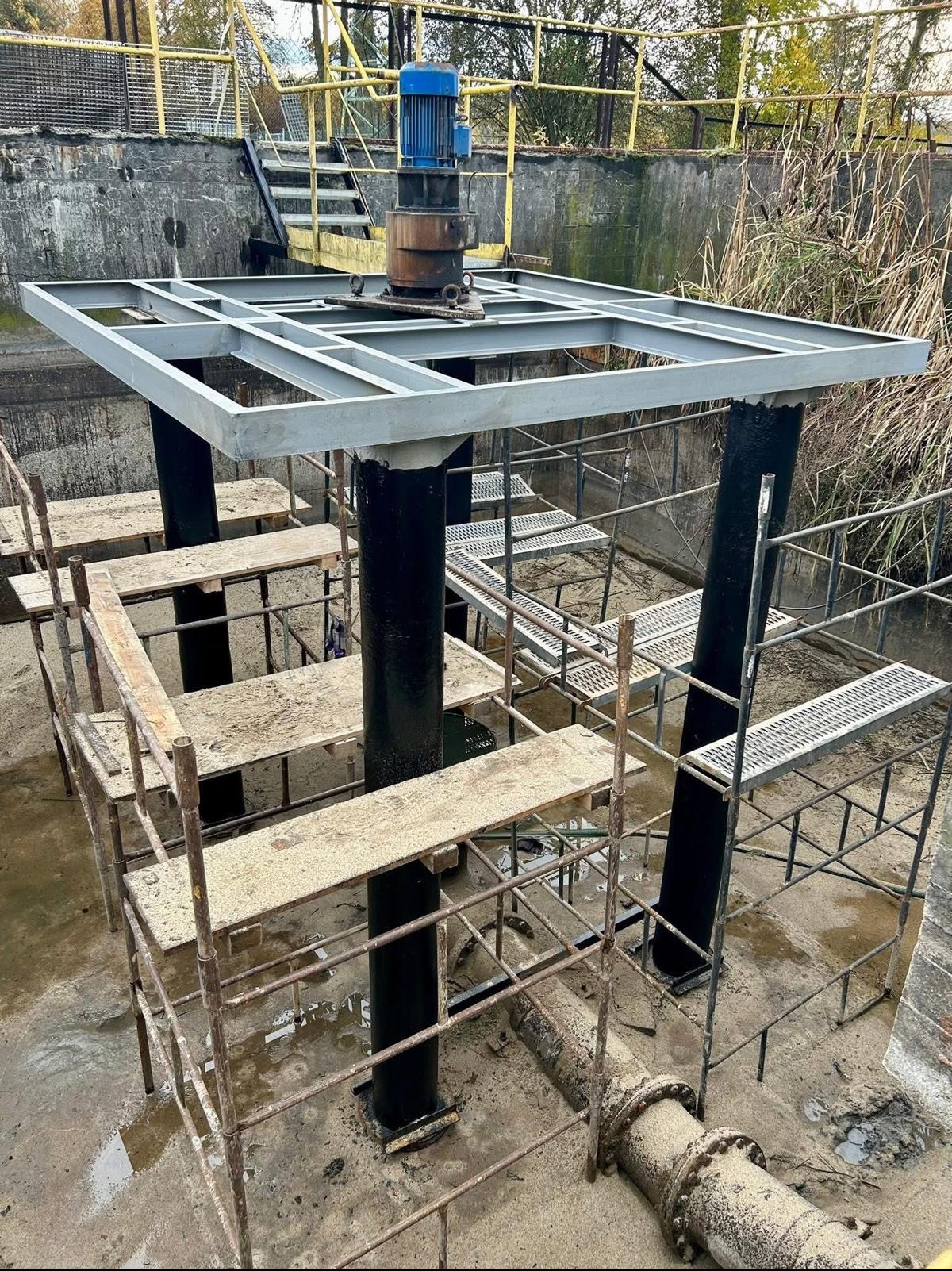Konstrukcja stalowa z silnikiem w oczyszczalni ścieków, otoczona rusztowaniami. Widoczne rury i betonowe elementy. Prace konserwacyjne lub budowlane w toku.