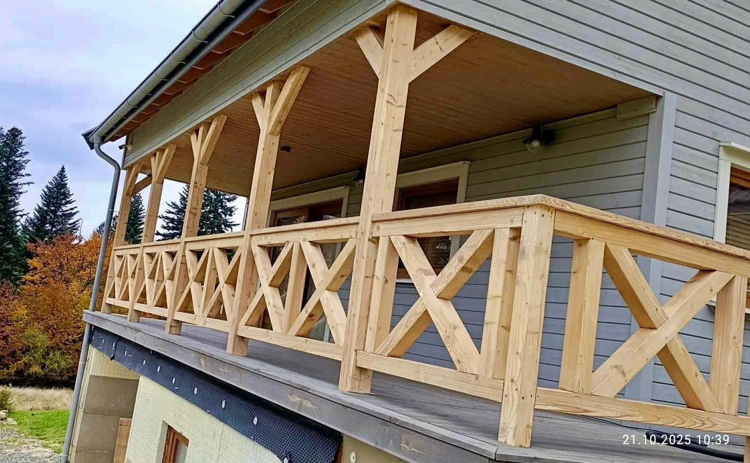 Drewniany balkon z balustradą w kształcie krzyży, wsparty na solidnych słupach. Szare ściany domu w tle. Widok na las i łąkę. Elementy konstrukcyjne z surowego drewna.