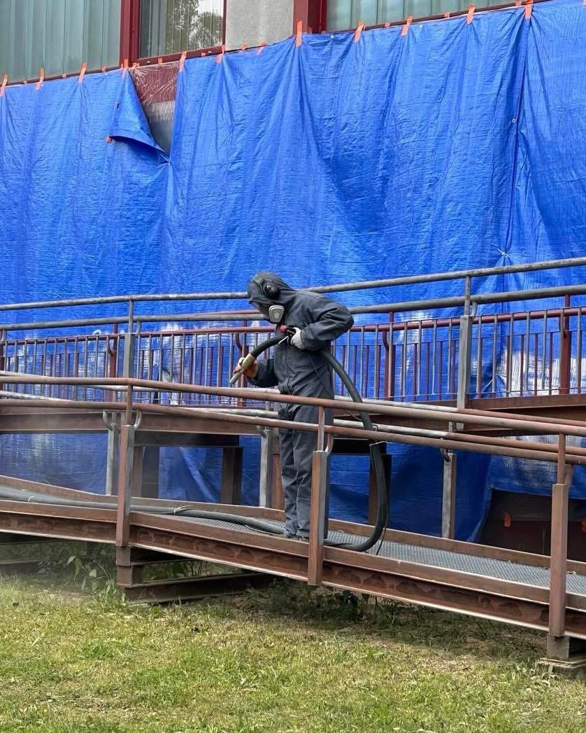 Pracownik w kombinezonie ochronnym piaskuje metalową konstrukcję na zewnątrz budynku, zabezpieczonego niebieską plandeką. Widoczny pył i trawa.