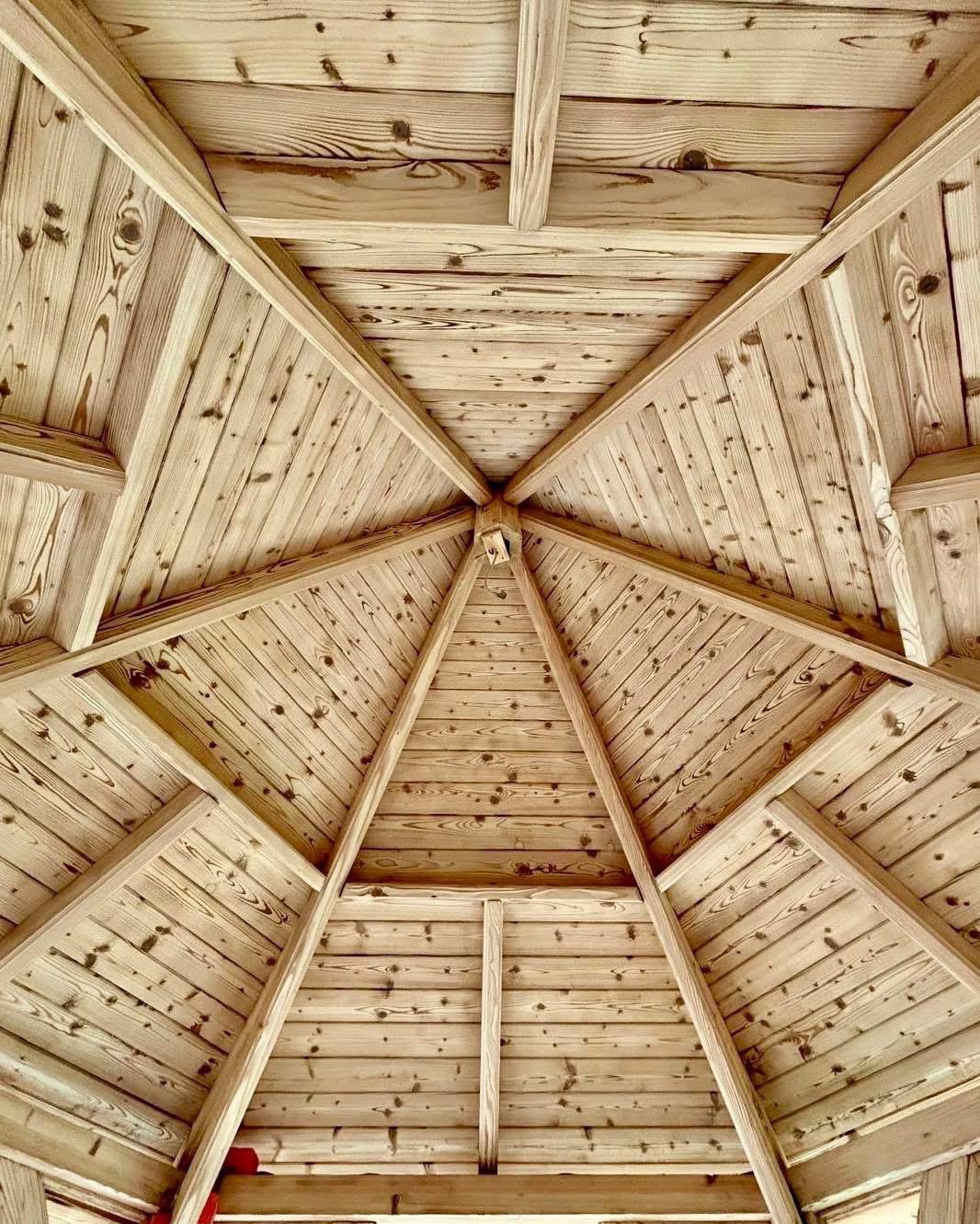 Drewniana konstrukcja altany z widocznym wzorem słojów, jasne drewno, symetryczny układ belek i desek, widok z dołu, centralny punkt zbiegu.