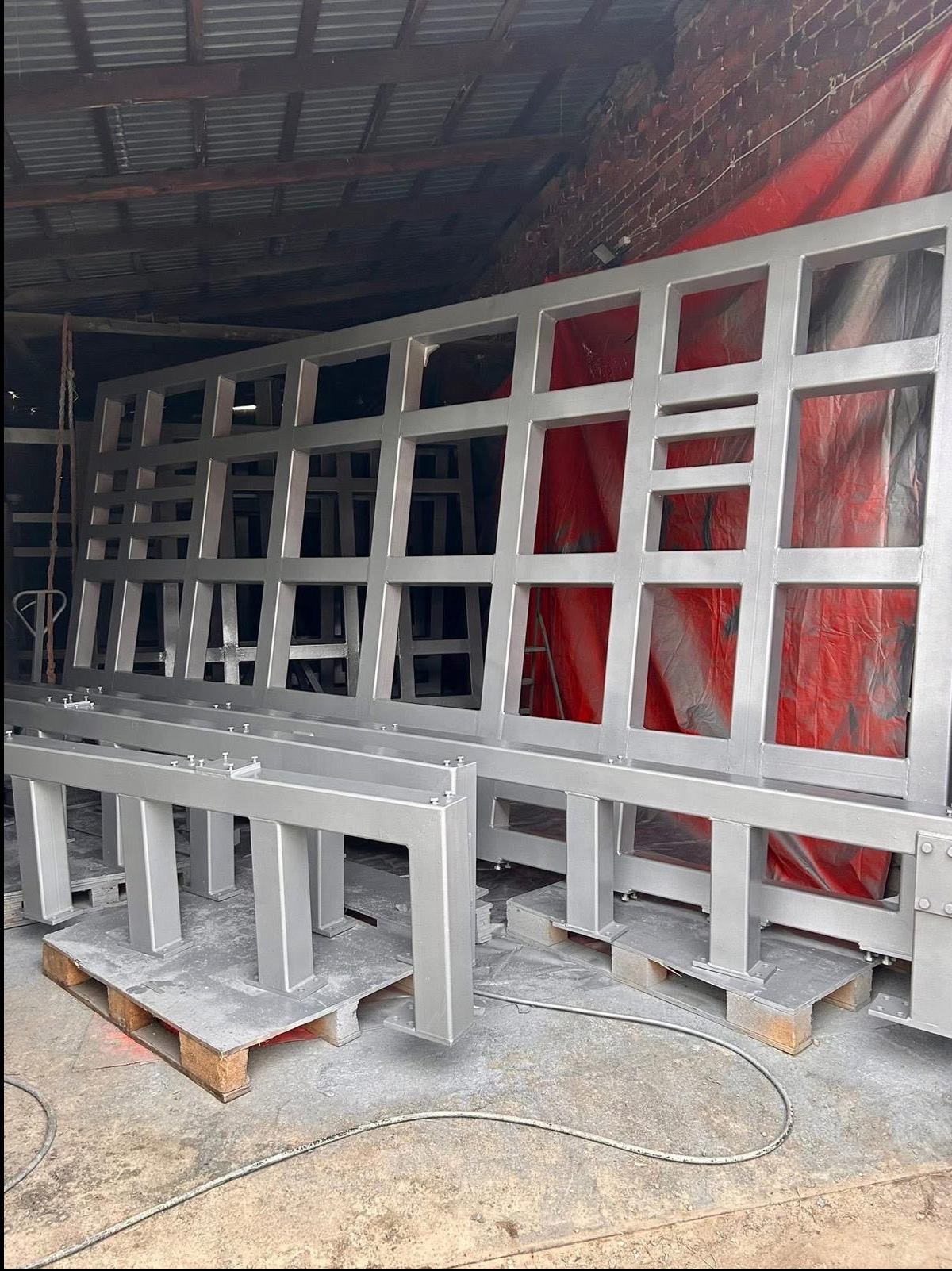 Metalowa konstrukcja kratowa i podpory po piaskowaniu i malowaniu na srebrno, ustawione w hali na paletach. Widoczna ceglana ściana i czerwona plandeka w tle.