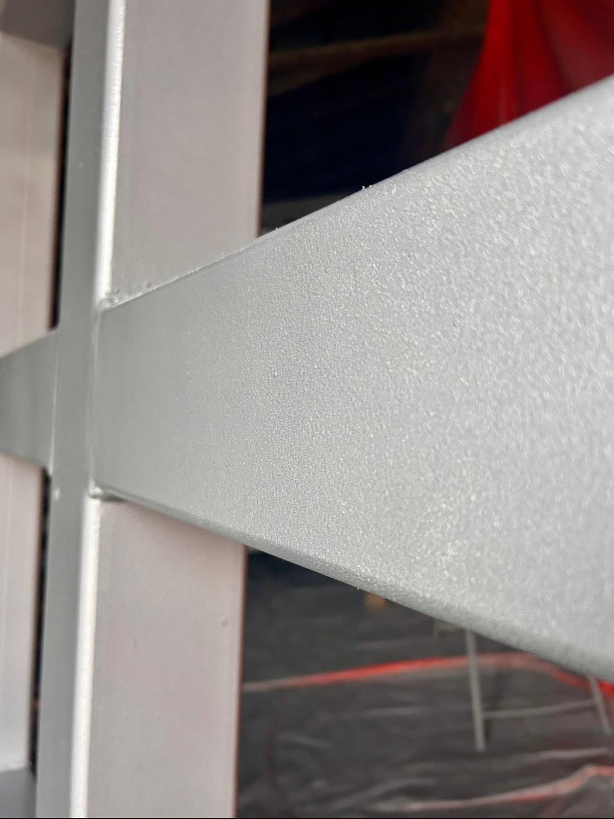 Metalowa konstrukcja po obróbce, pokryta jednolitą, srebrzystą powłoką. Widoczna faktura powierzchni i detale łączeń. Tło częściowo rozmyte, z elementami czerwieni.