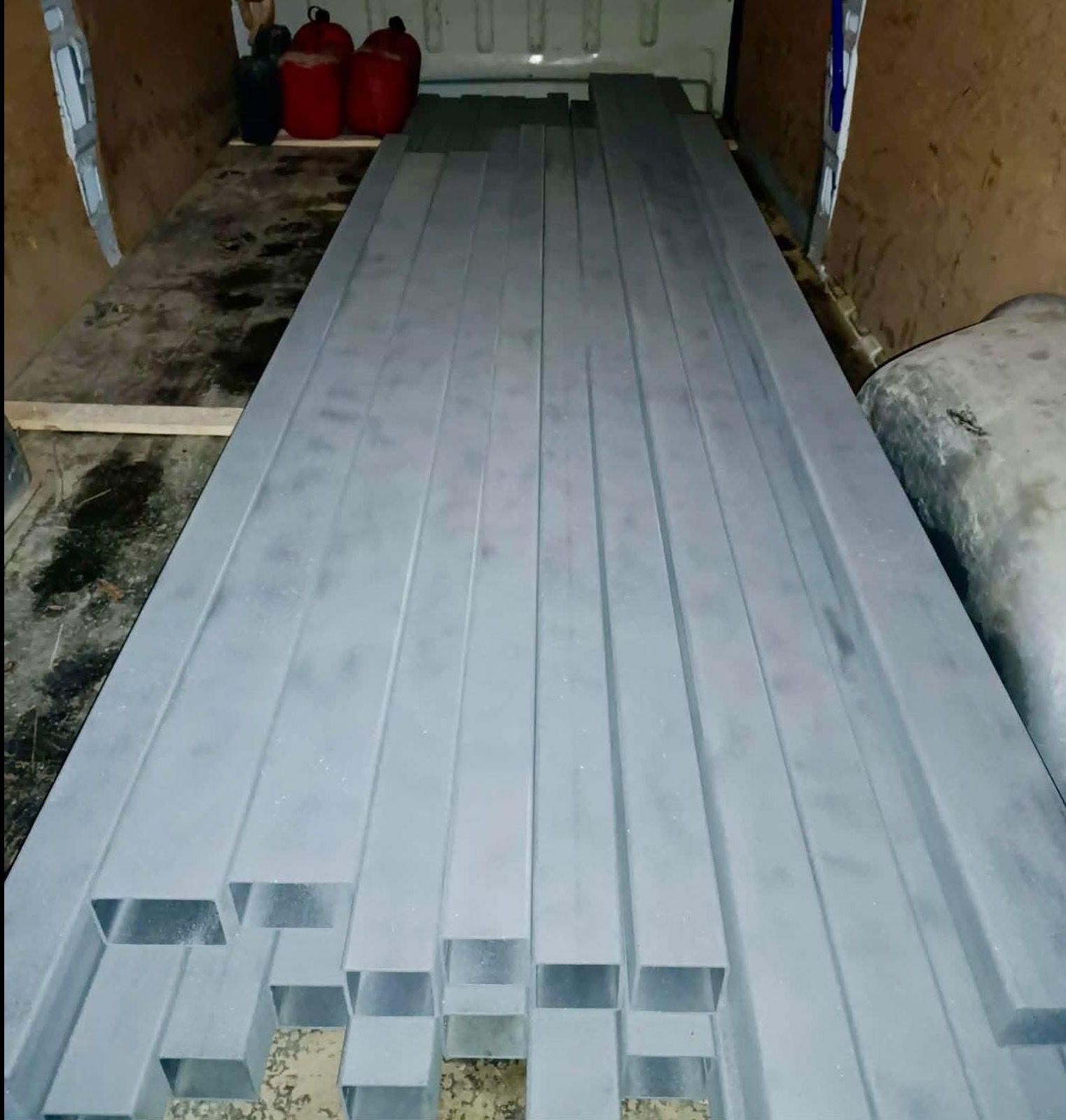 Szare, metalowe profile o przekroju kwadratowym, ułożone wzdłużnie w przestrzeni ładunkowej pojazdu. Widoczne ślady użytkowania i pojemniki w tle.