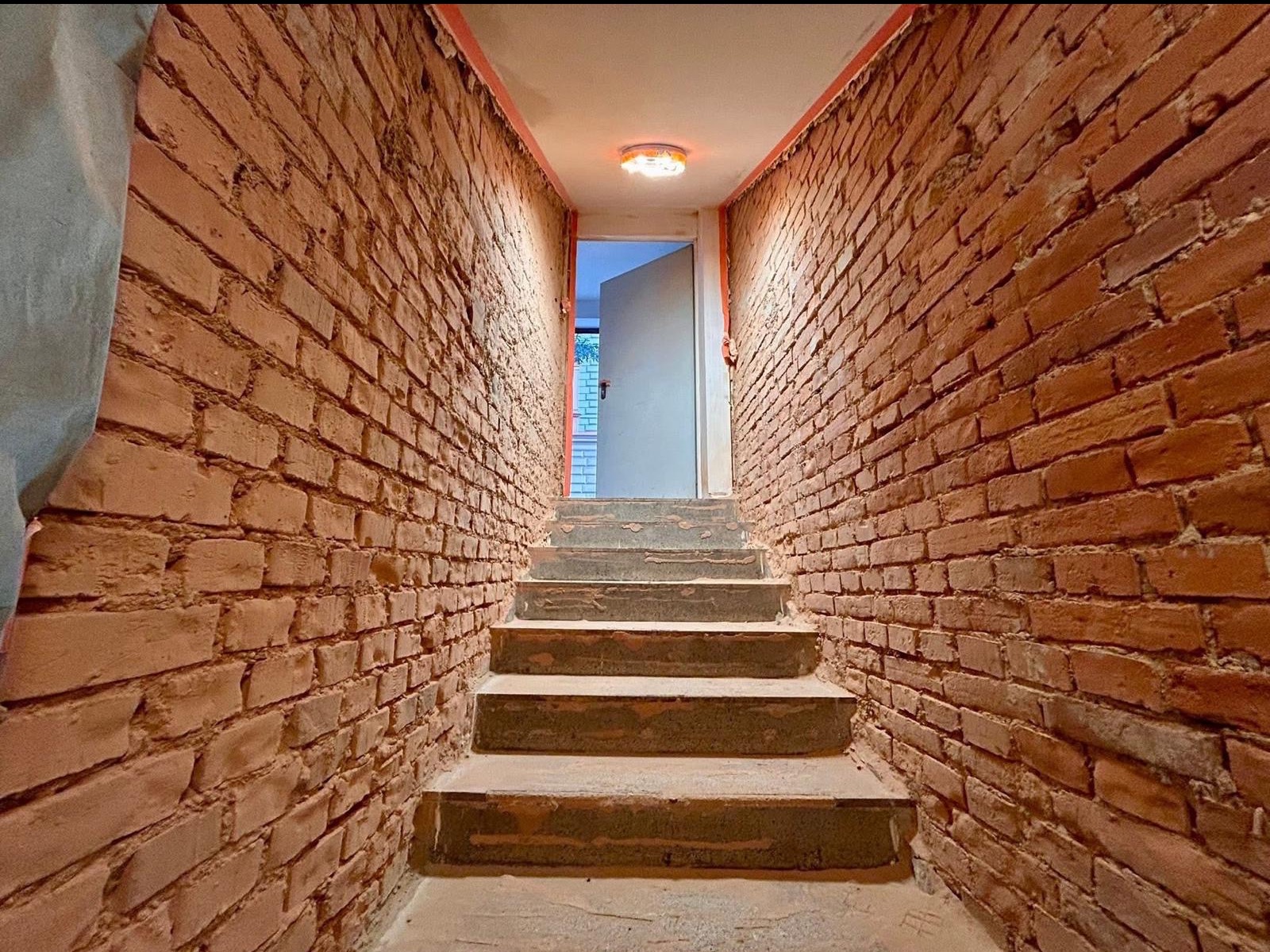 Ceglana ściana po obróbce, widoczne schody i otwarte drzwi. Surowe wykończenie, perspektywa z dołu, ciepłe oświetlenie. Detal ściany z cegły.