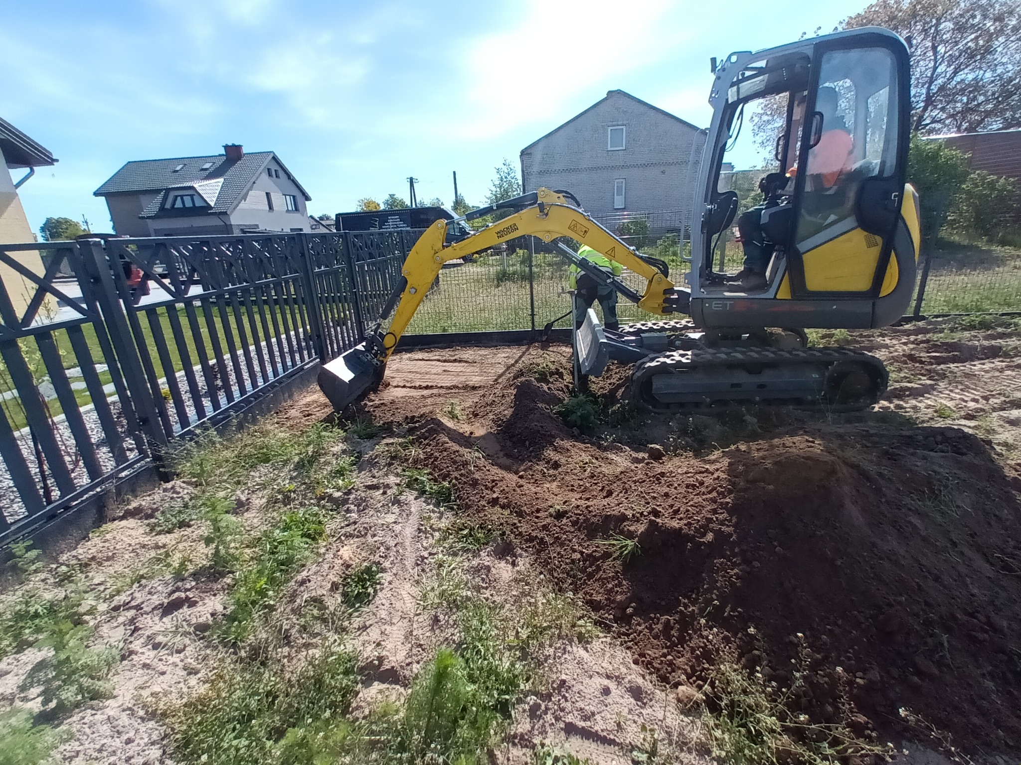 Żółta mini koparka Wacker Neuson podczas prac ziemnych przy ogrodzeniu posesji. Widoczny operator w kabinie i wykopany rów w ziemi. Prace przygotowawcze pod kabel.