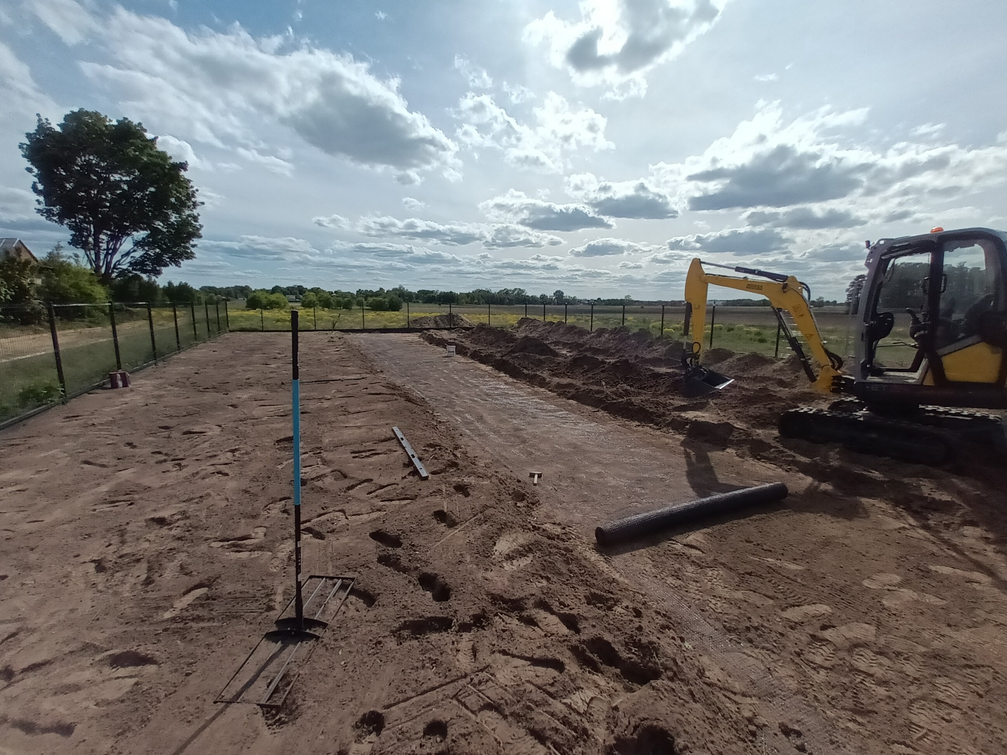Przygotowanie terenu pod instalacje: wykop, mini koparka, narzędzia. Widok na ogrodzony teren z drzewem i zachmurzonym niebem w tle. Prace ziemne w toku.