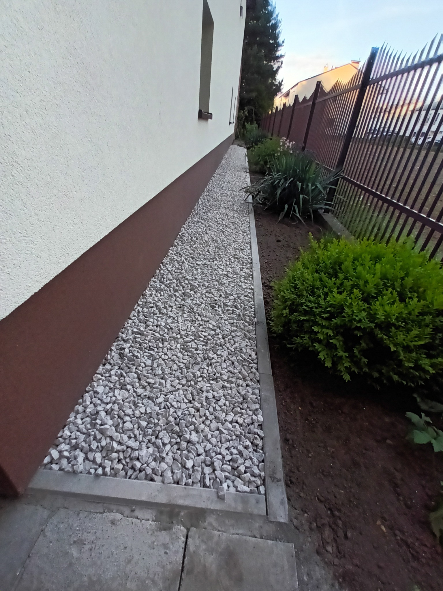 Ścieżka z białego grysu oddzielona obrzeżem od rabaty z roślinami, biegnąca wzdłuż elewacji budynku i ogrodzenia z metalowych prętów.