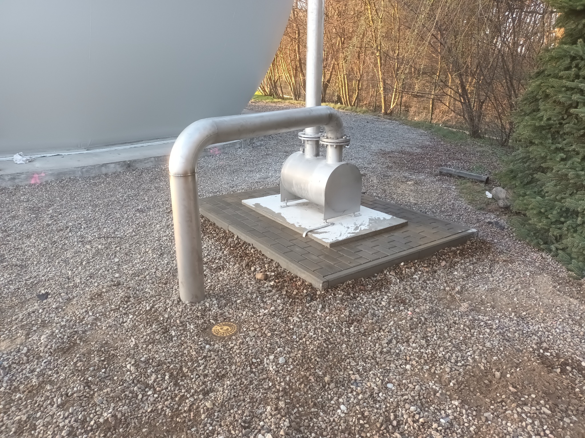 Metalowa instalacja telekomunikacyjna na zewnątrz, w tle drzewa i część zbiornika. Widoczny właz rewizyjny w ziemi. Instalacja na betonowej podstawie.