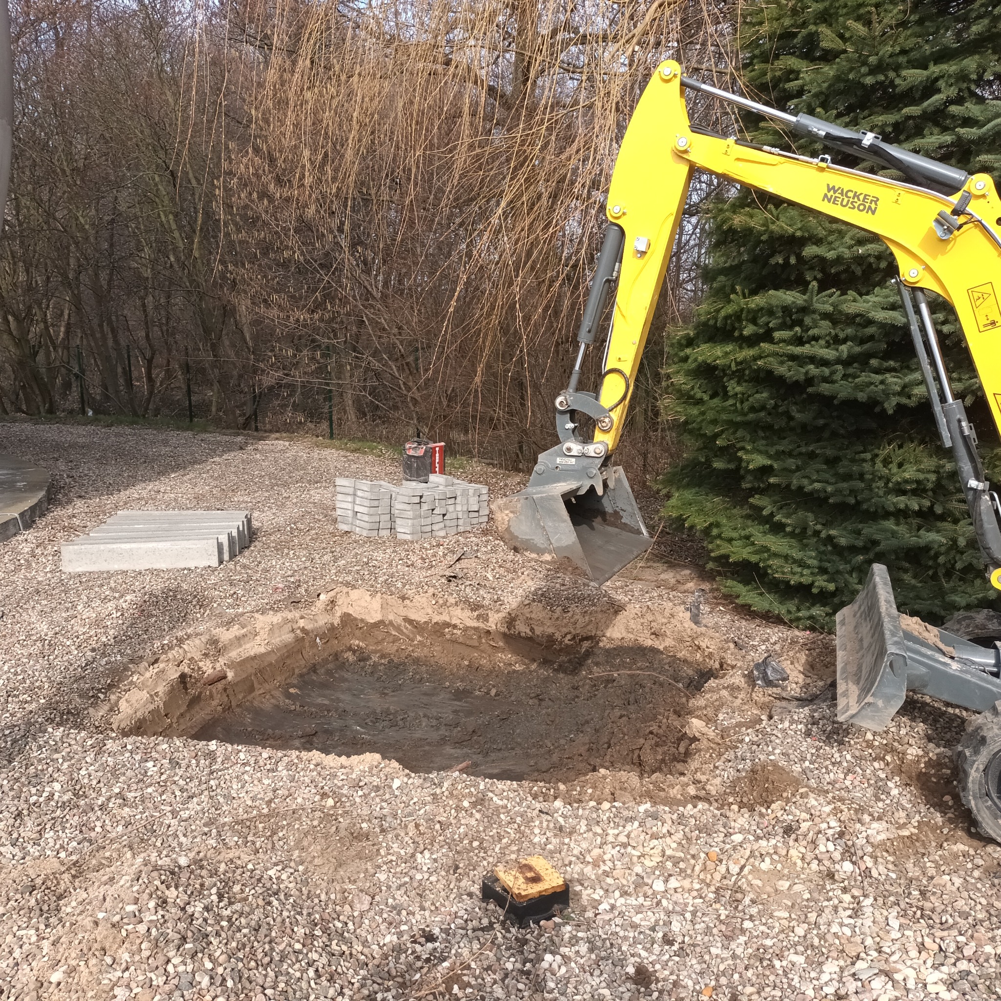 Żółta mini koparka Wacker Neuson w trakcie wykopu pod instalację telekomunikacyjną; widoczne ułożone płyty chodnikowe i kostka brukowa.