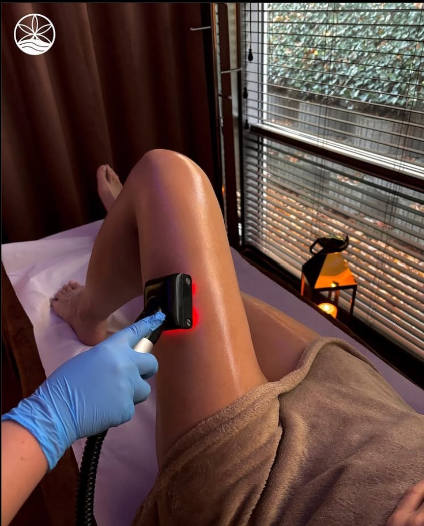 Zabieg na nogę z użyciem urządzenia emitującego czerwone światło, wykonywany przez osobę w niebieskich rękawiczkach, widok z góry na leżącą osobę na białym prześcieradle w salonie.