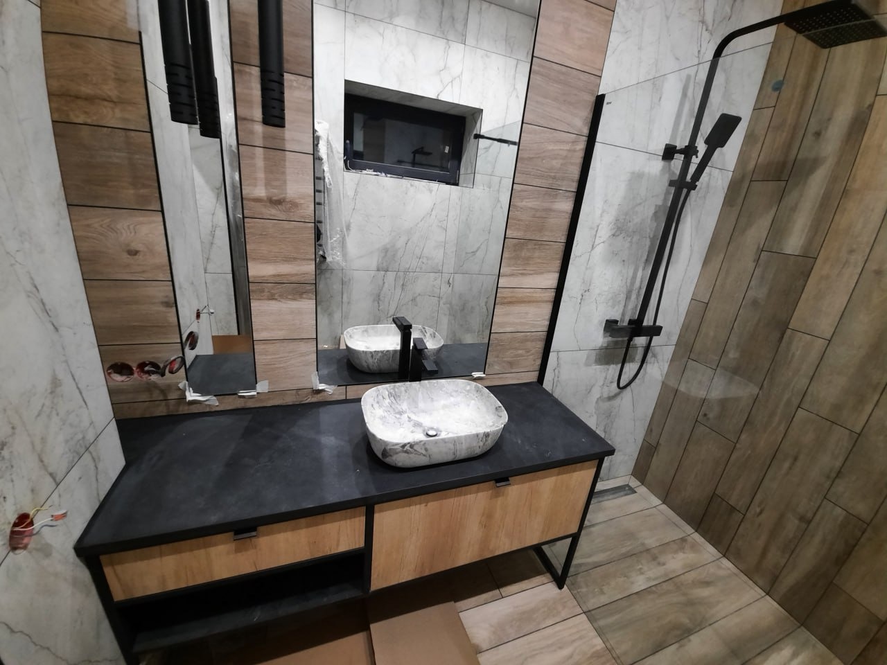 Nowoczesna łazienka z umywalką nablatową w marmurkowy wzór, czarną armaturą i płytkami imitującymi drewno oraz marmur.