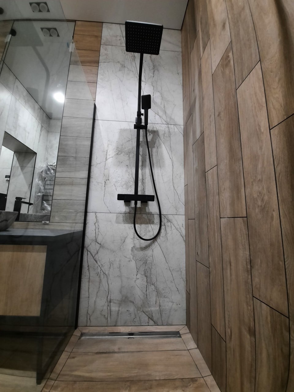 Nowoczesna kabina prysznicowa z czarną armaturą i odpływem liniowym, wykończona płytkami imitującymi marmur i drewno.