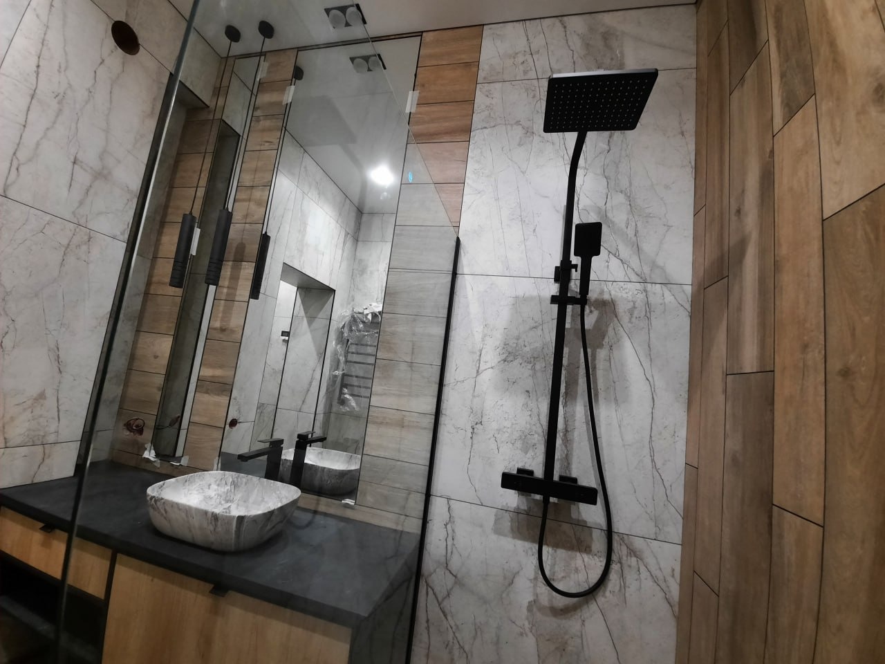 Nowoczesna łazienka z marmurowymi płytkami, czarną armaturą i umywalką w stylu kamiennym, oświetlenie punktowe i lustro z podłużnymi lampami.