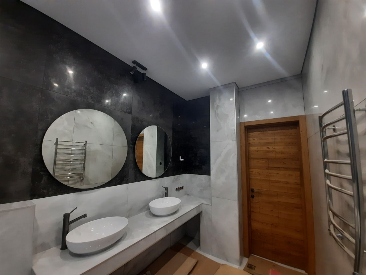 Nowoczesna łazienka z dwoma okrągłymi lustrami, podwójną umywalką nablatową, czarną armaturą i grzejnikiem drabinkowym, wykończona płytkami w odcieniach szarości i bieli.