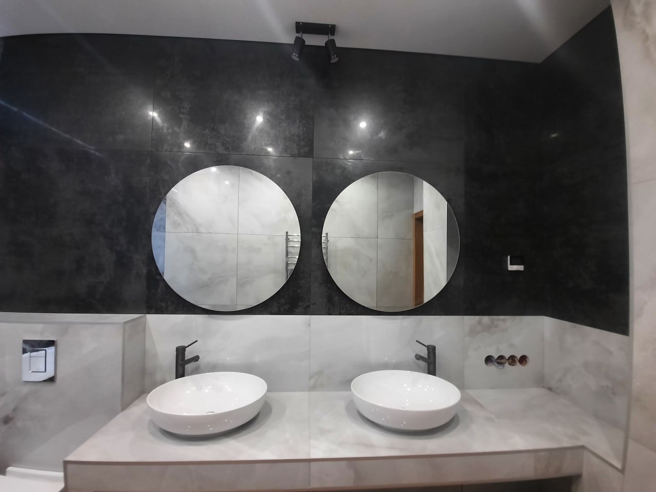 Nowoczesna łazienka z dwoma okrągłymi lustrami nad umywalkami wpuszczonymi w marmurowy blat, czarne baterie i ciemna, błyszcząca ściana w tle.
