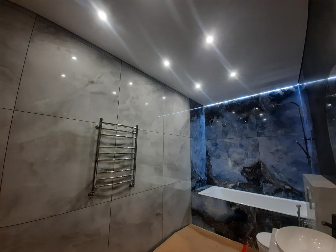 Wykończona łazienka z płytkami imitującymi marmur w odcieniach szarości i błękitu, chromowany grzejnik drabinkowy na ścianie, podświetlenie LED przy suficie.