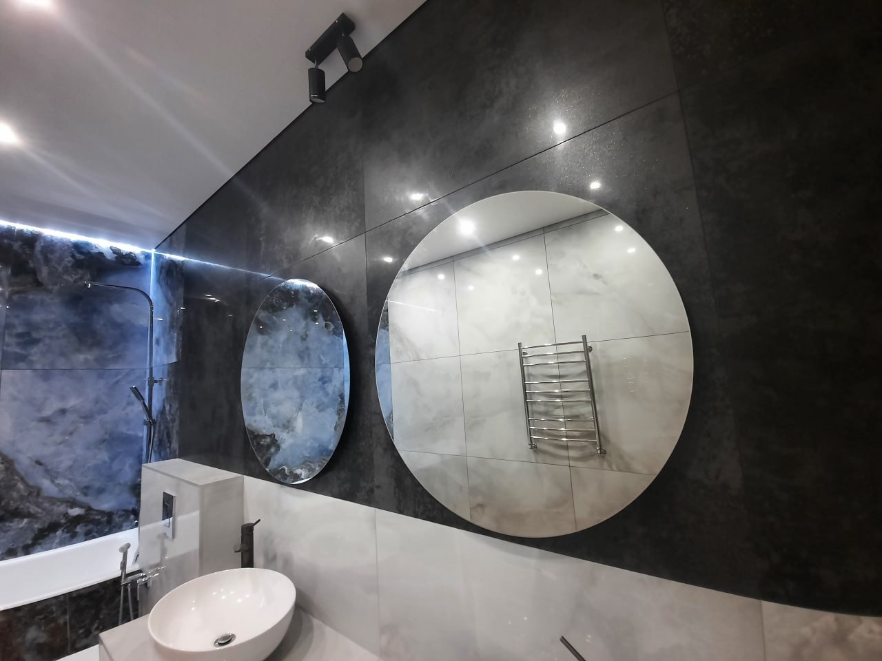 Nowoczesna łazienka z czarno-białymi płytkami, okrągłymi lustrami i chromowanym grzejnikiem drabinkowym odbitym w lustrze, oświetlenie punktowe na suficie.