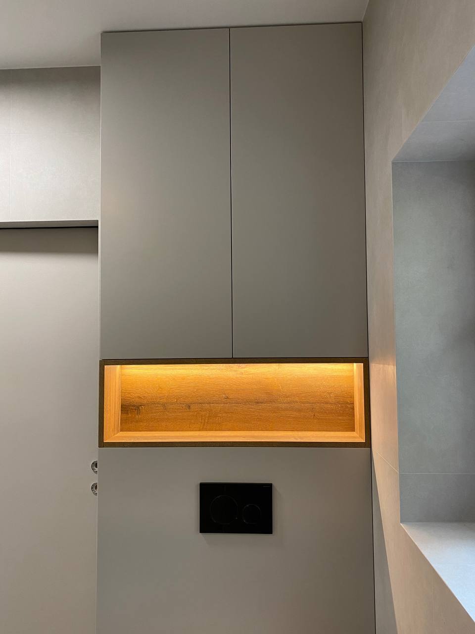 Szara zabudowa meblowa w łazience z podświetlaną drewnianą wnęką nad czarnym przyciskiem spłuczki WC.